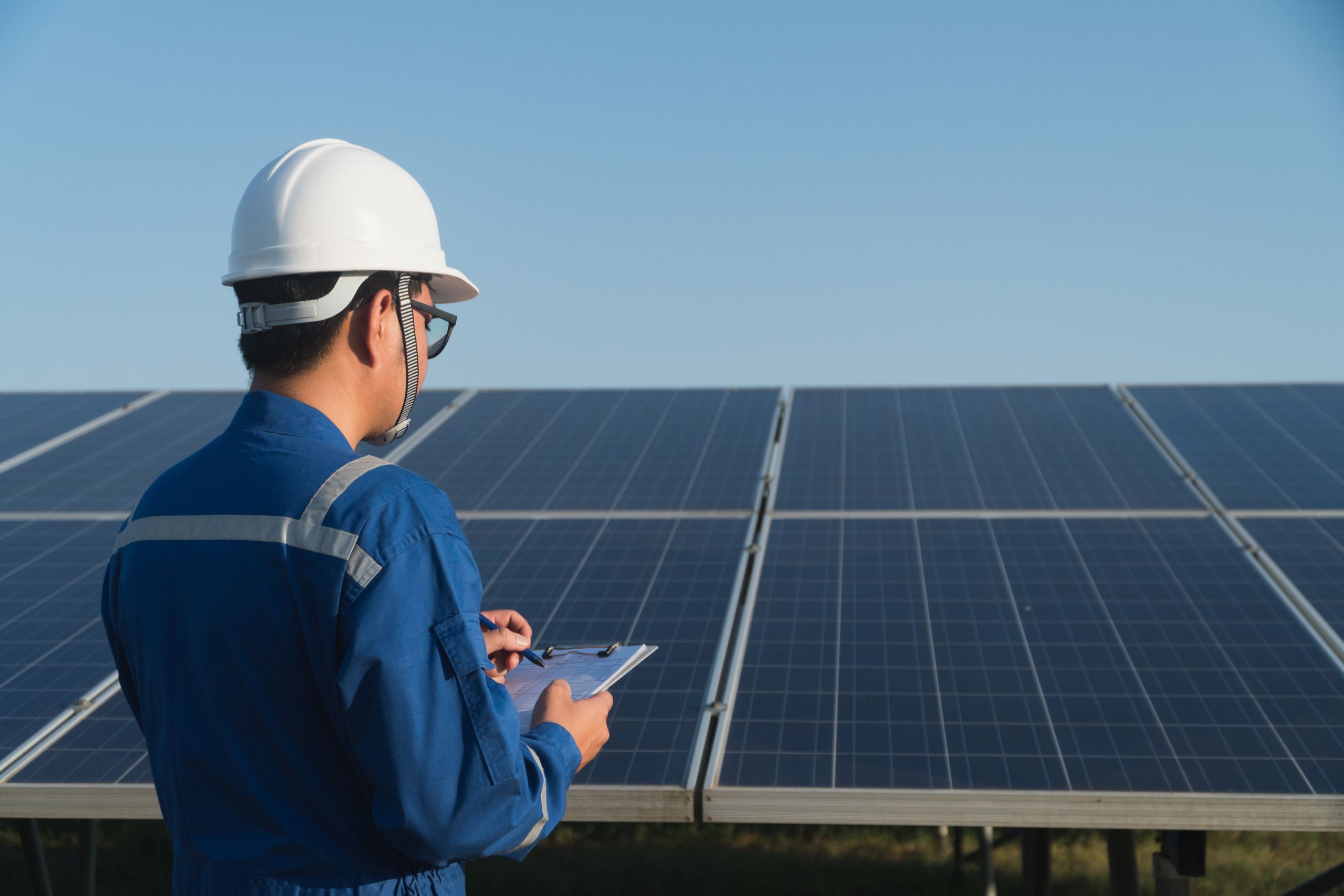 A solar panel installer wearing a hard hat, standing in front of a roof with solar panels